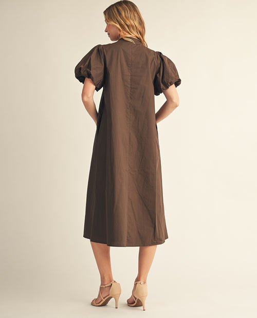 A woman shown from the back wearing a brown midi dress with puffed short sleeves and beige high-heeled sandals, standing against a plain light beige studio background.