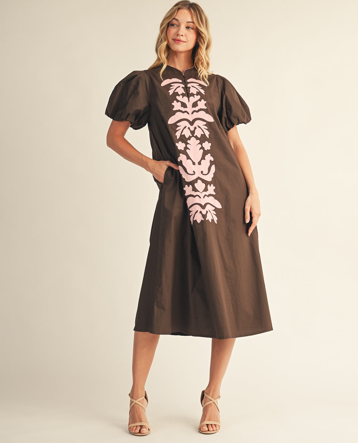 A woman modeling a brown midi dress with puff sleeves and a pink damask-style vertical pattern on the front, photographed against a plain light beige studio background.