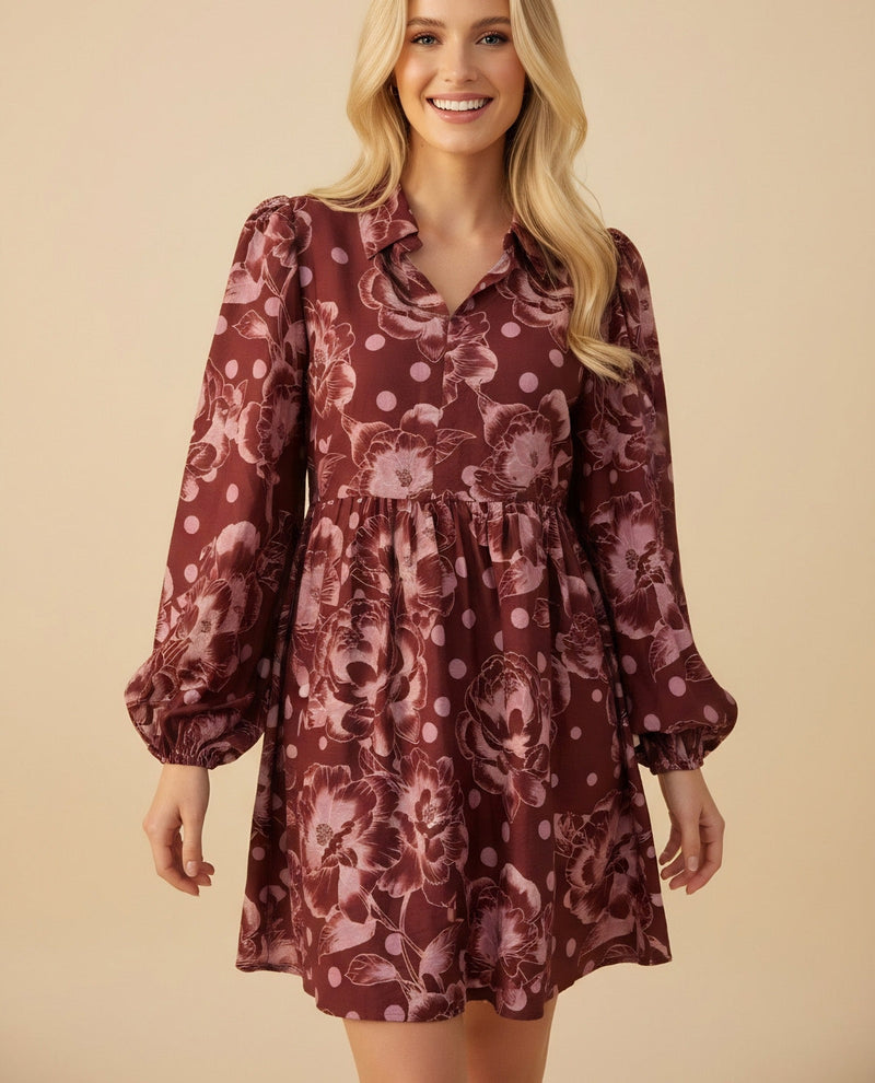 A smiling blonde woman models a long-sleeve mini dress with a floral and polka dot pattern in dusty rose and burgundy, set against a solid beige studio background.