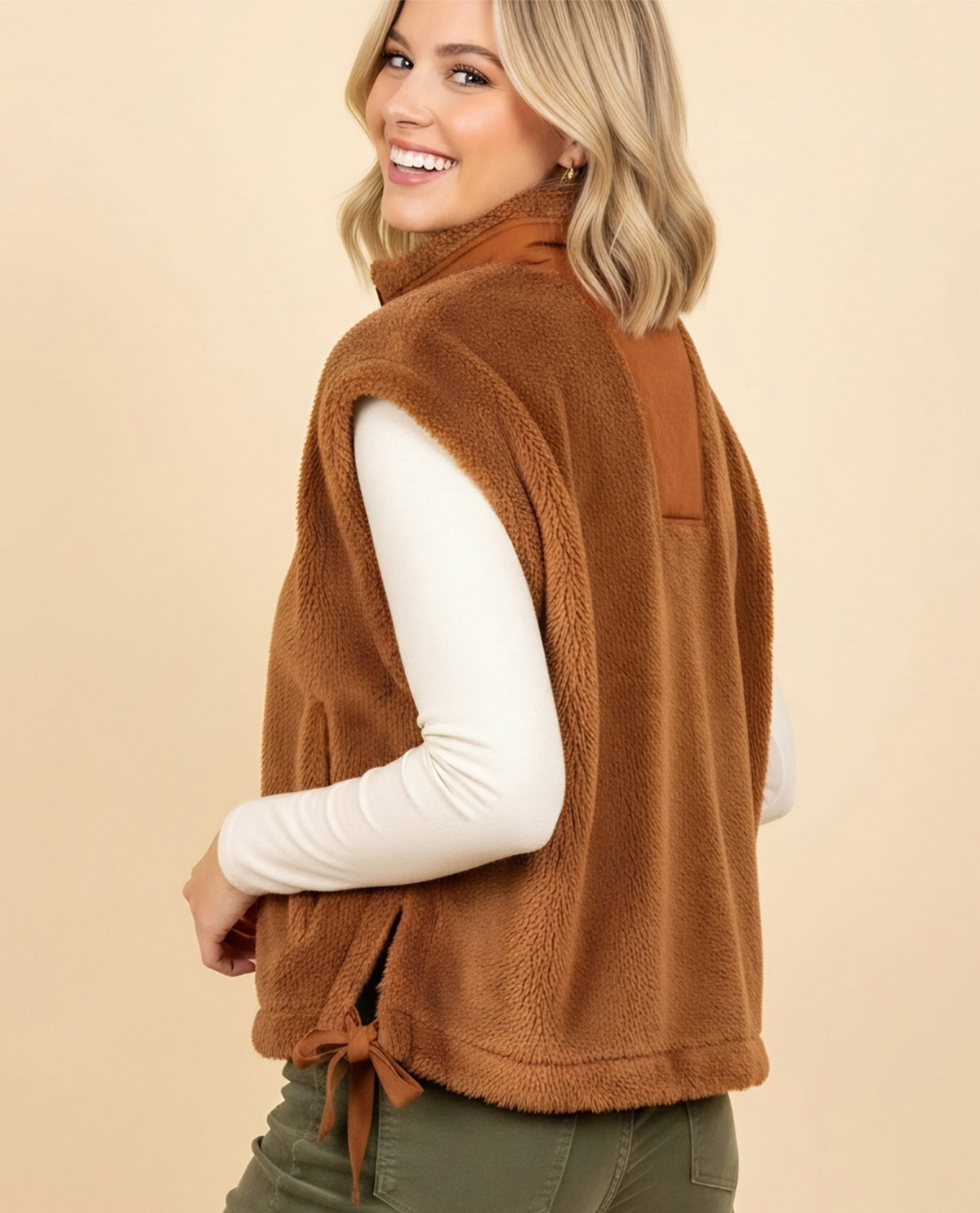 A woman wearing a brown sherpa fleece vest over a white long-sleeve top and olive green pants, shown from a side-back angle against a solid beige studio background.