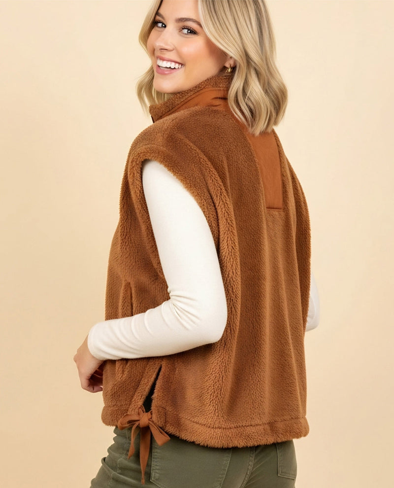 A woman wearing a brown sherpa fleece vest over a white long-sleeve top and olive green pants, shown from a side-back angle against a solid beige studio background.