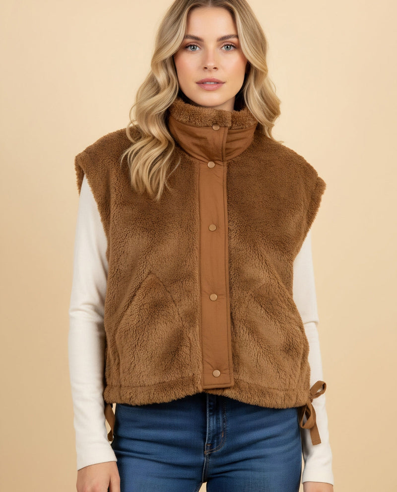 A woman wearing a brown faux fur sleeveless vest over a white long-sleeve shirt and blue jeans, standing against a solid beige background.