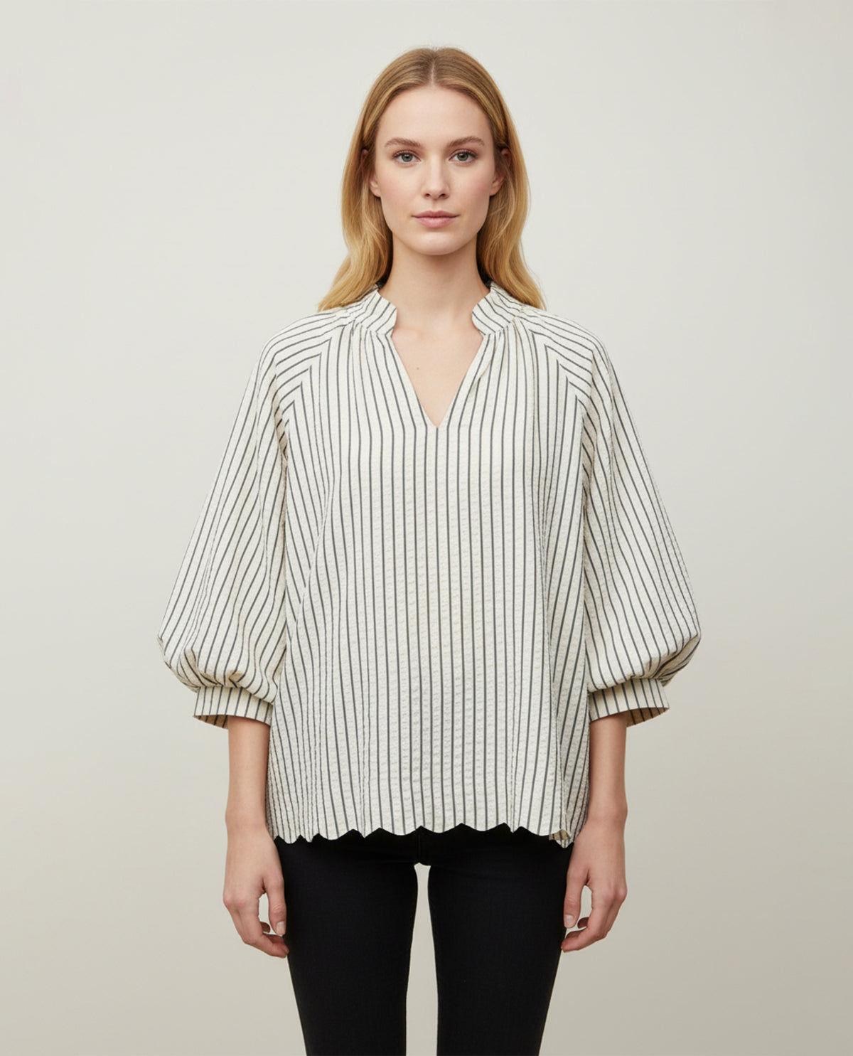 A woman modeling a cream-colored blouse with thin black vertical stripes, featuring a split collar, a split V-neckline, and voluminous elbow-length puff sleeves. She is also wearing black pants, set against a plain light gray studio background.
