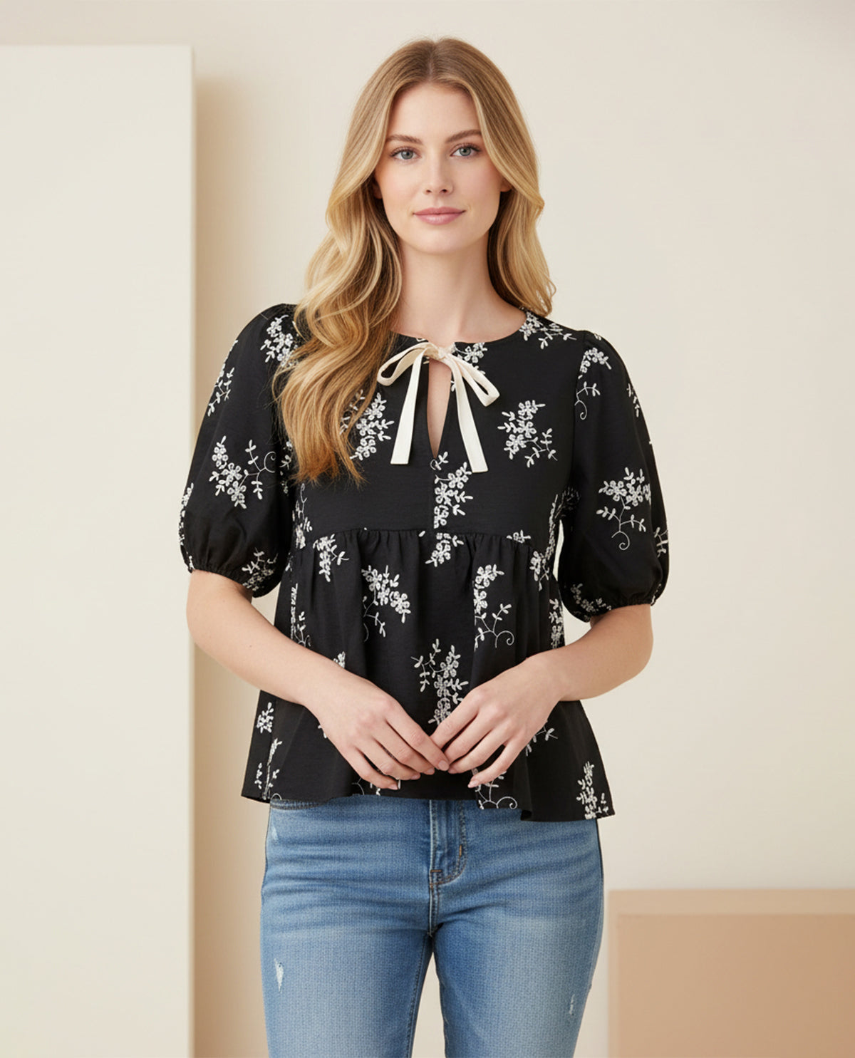 Woman wearing a black floral blouse with a white ribbon and blue jeans against a neutral background
