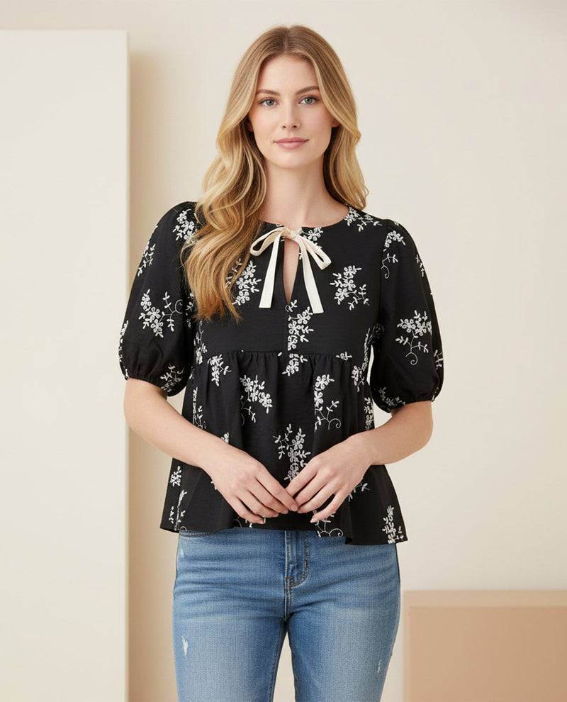 Woman wearing a black floral blouse with a white ribbon and blue jeans against a neutral background