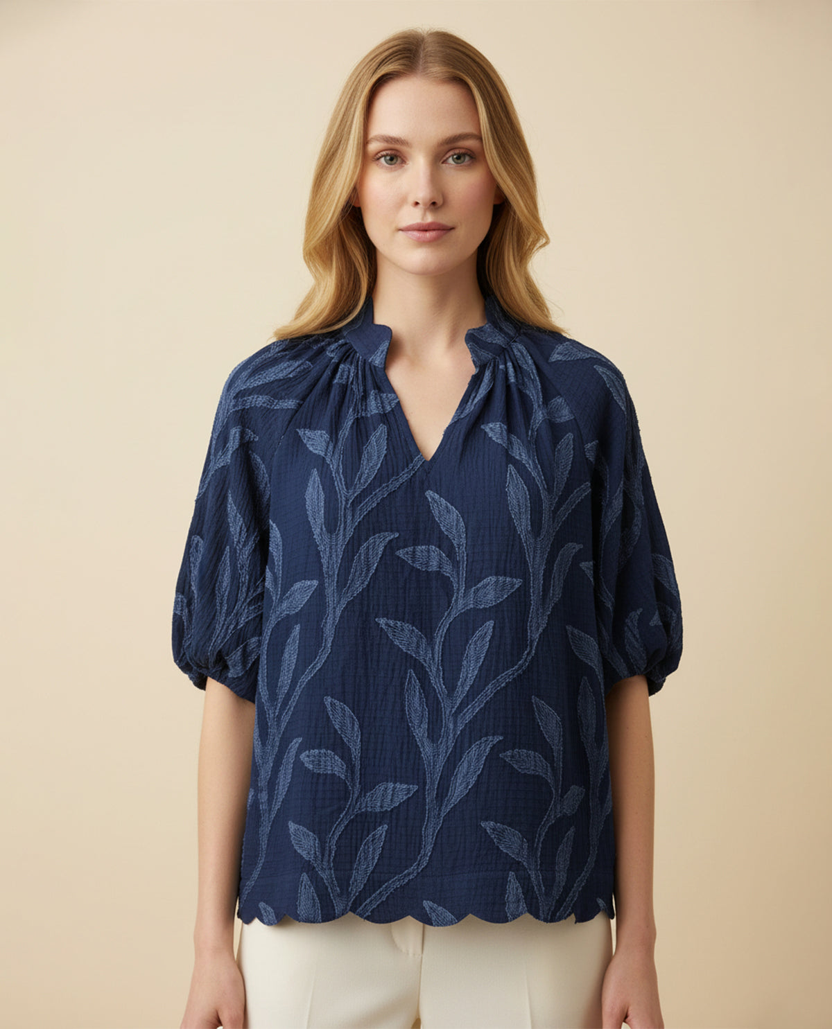 A woman wearing a navy blue textured blouse with a tonal leaf pattern, short puff sleeves, and a split V-neckline, paired with cream pants against a solid beige background.