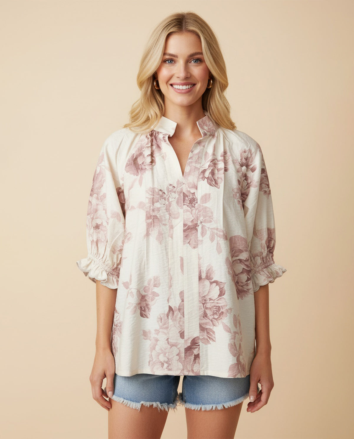 A smiling blonde woman models a loose white button-up blouse with a dusty pink floral print, ruffled cuffs, and a tie detail at the collar, paired with denim shorts against a solid beige background.