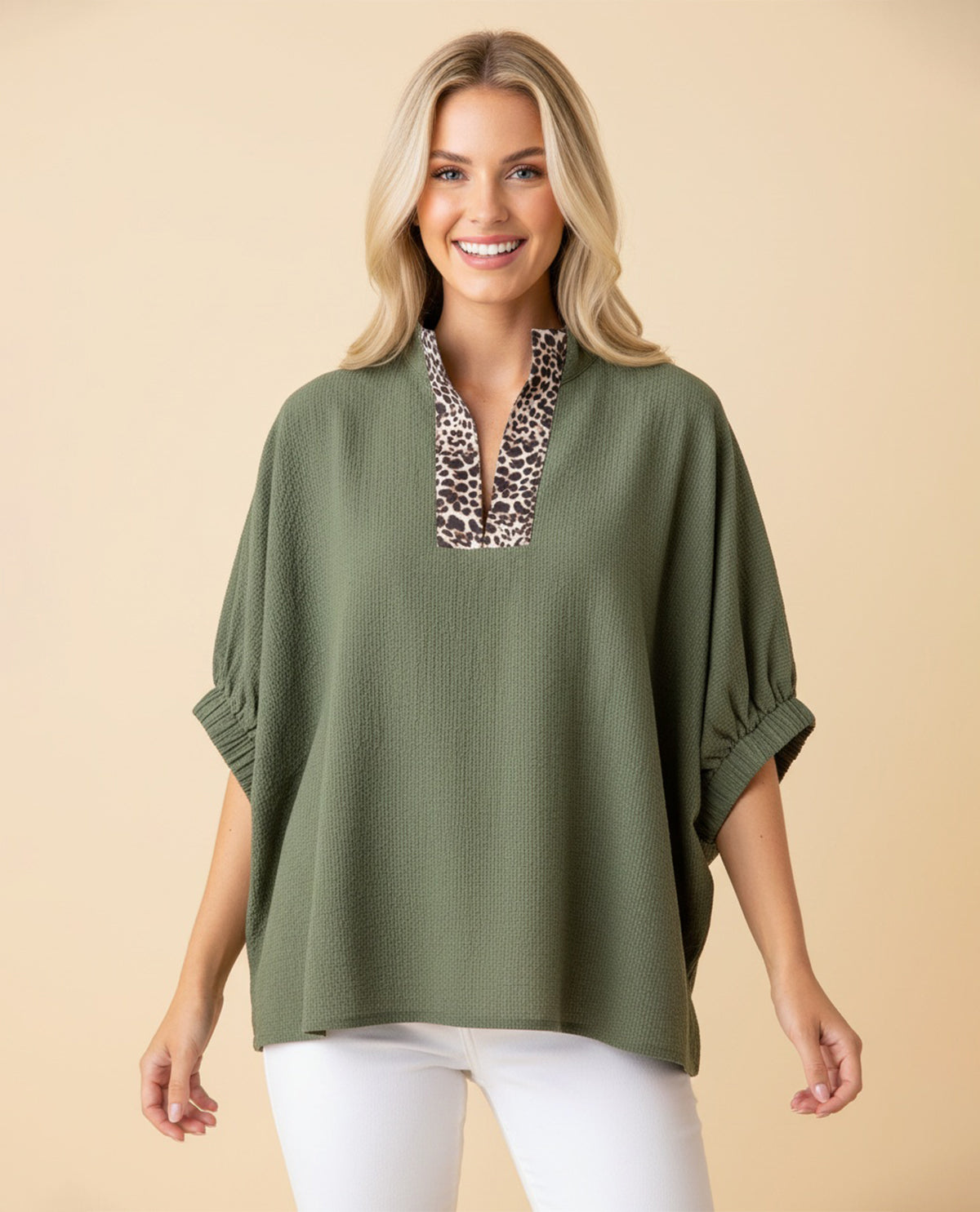 A woman modeling a loose olive green top with a leopard-print V-neck trim, paired with white pants, against a solid beige background.