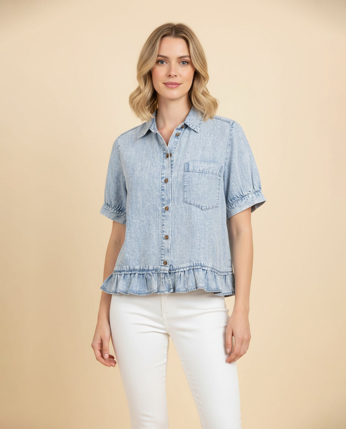 A woman modeling a light blue denim short-sleeve button-up shirt with a ruffled hem and chest pocket, paired with white pants, against a solid beige background.