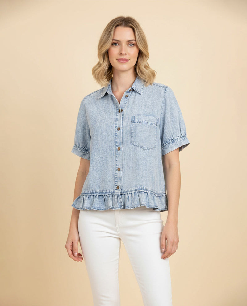 A woman modeling a light blue denim short-sleeve button-up shirt with a ruffled hem and chest pocket, paired with white pants, against a solid beige background.