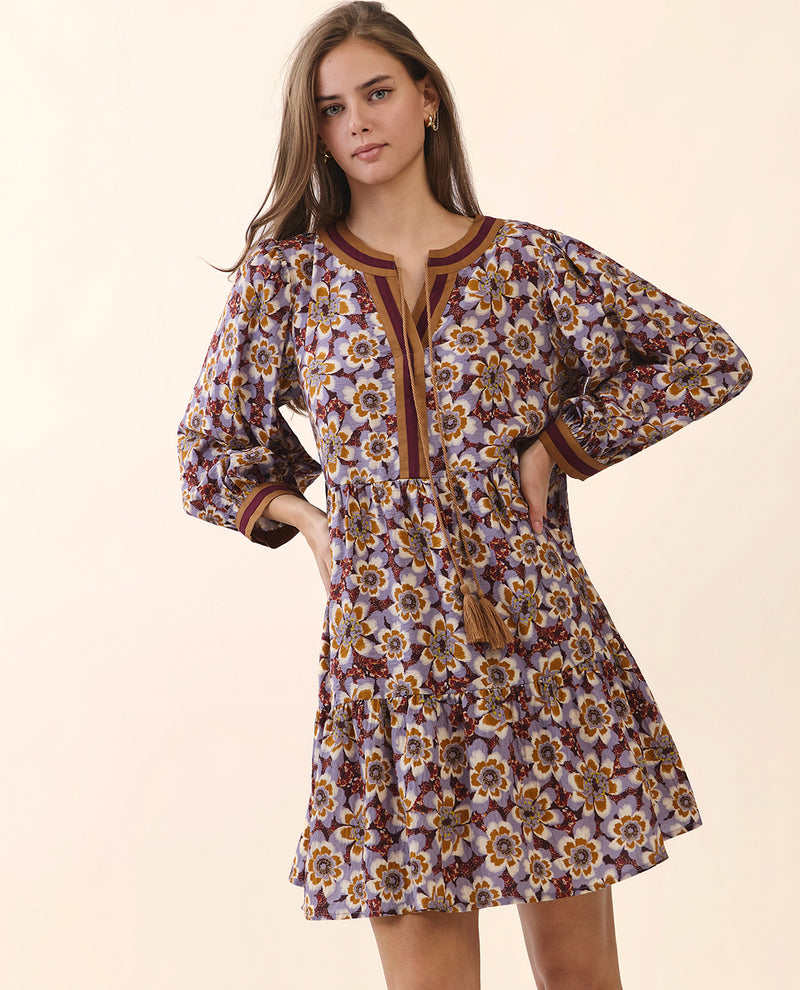 A woman modeling a long-sleeve bohemian-style mini dress with a purple, brown, and yellow floral pattern, set against a solid beige background.