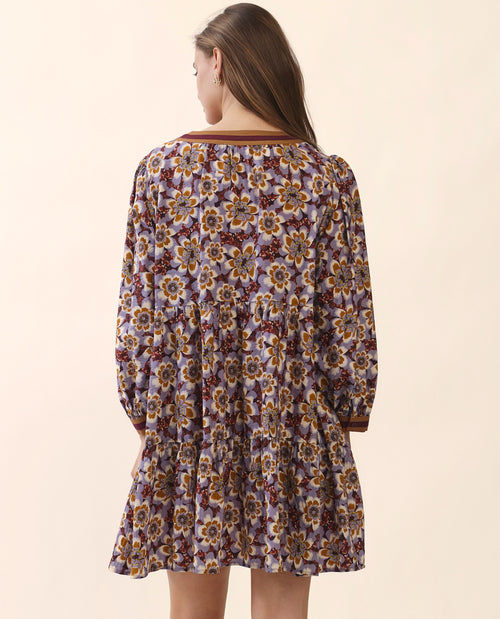A woman viewed from the back wearing a long-sleeve tiered dress with an all-over floral pattern in purple, yellow, and brown tones, set against a solid light beige background.