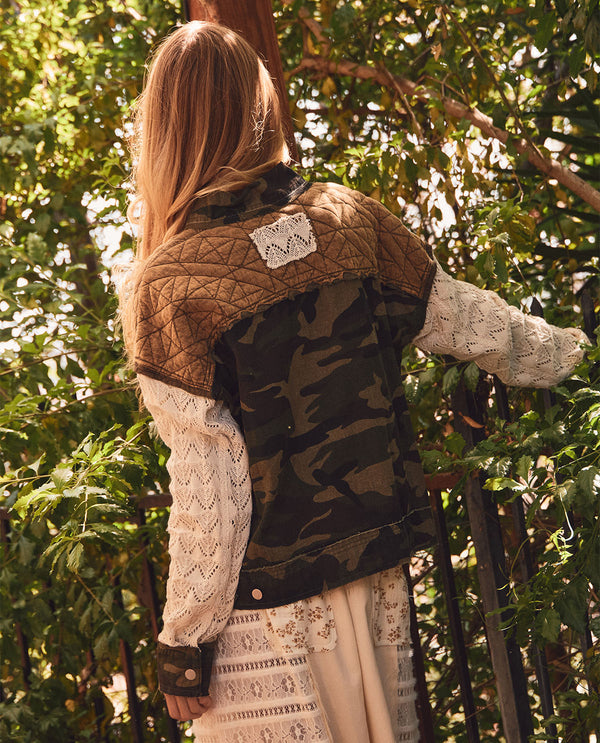 A woman viewed from behind wearing a quilted jacket with brown and camouflage panels over cream lace sleeves and a cream skirt, standing outdoors among green trees.