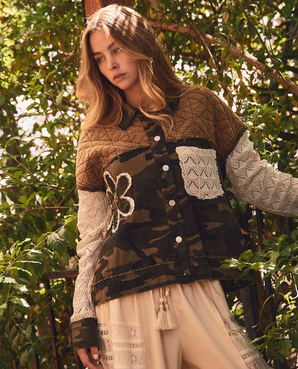 A woman wearing a bohemian-style patchwork jacket with quilted brown panels, a camouflage front pocket, and cream lace sleeves, standing outdoors in front of green foliage.