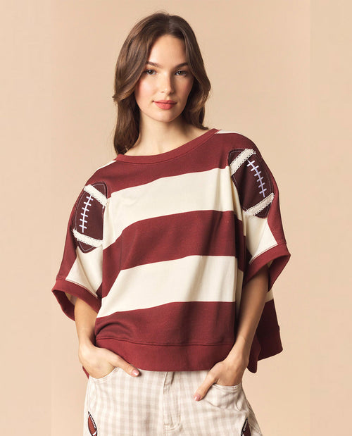 A woman modeling a loose-fitting, short-sleeve crewneck sweatshirt with horizontal burgundy and cream stripes and football graphic details on the shoulders, paired with light plaid pants against a solid beige background.