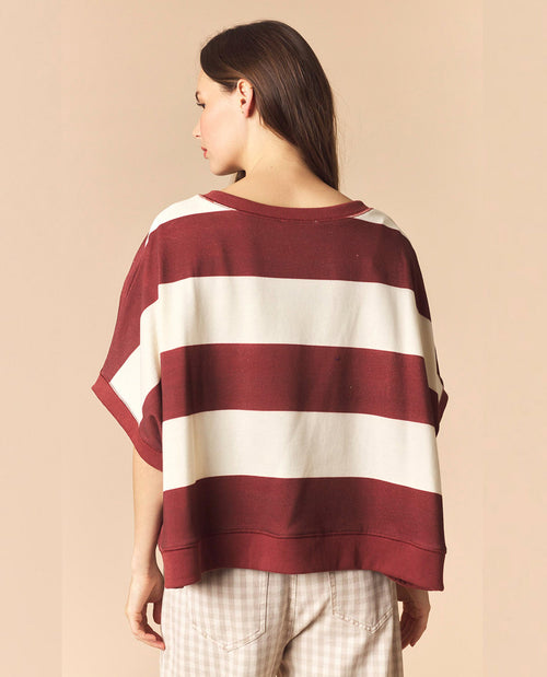 A woman shown from the back wearing a loose, short-sleeve top with wide horizontal stripes in burgundy and off-white, paired with light beige checkered pants, against a solid beige background.
