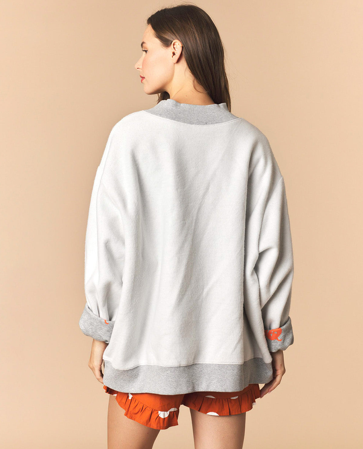 A woman shown from the back wearing an oversized light gray sweatshirt with contrasting medium gray ribbed collar, cuffs, and hem, paired with orange shorts, against a solid beige studio background.