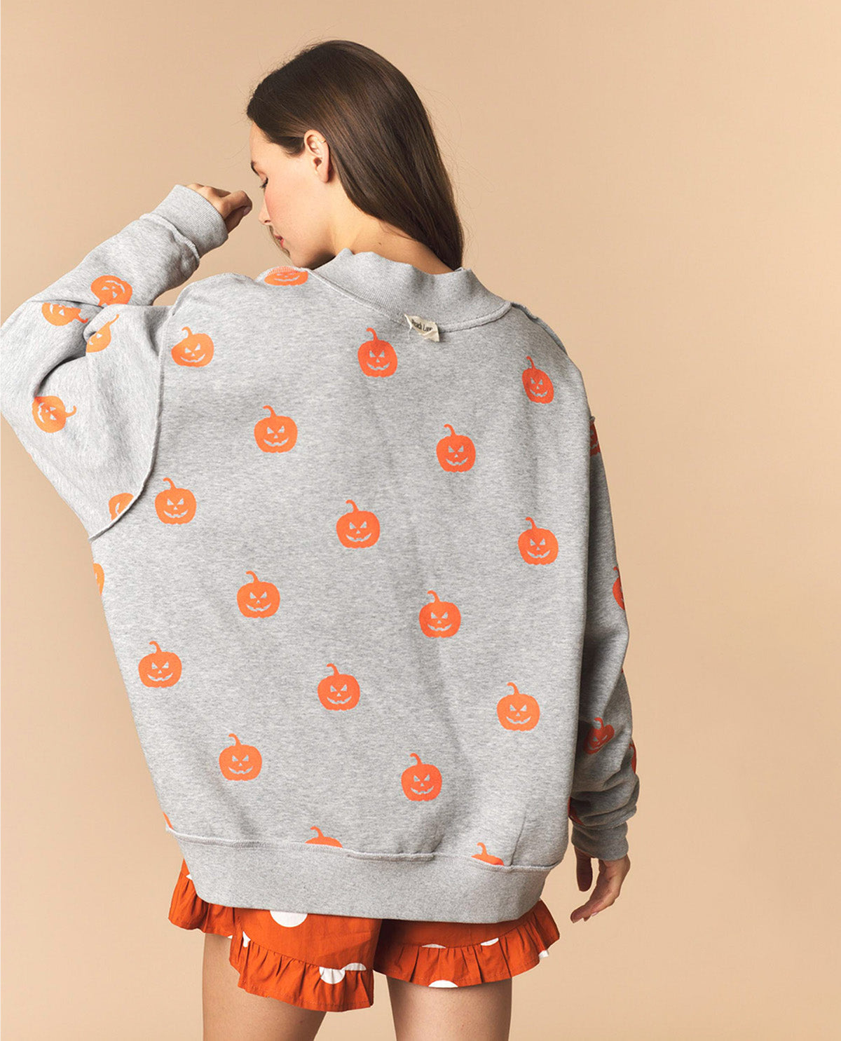 A woman shown from the back wearing a light gray heather sweatshirt with an all-over orange pumpkin print, paired with orange ruffled shorts, against a solid beige background.