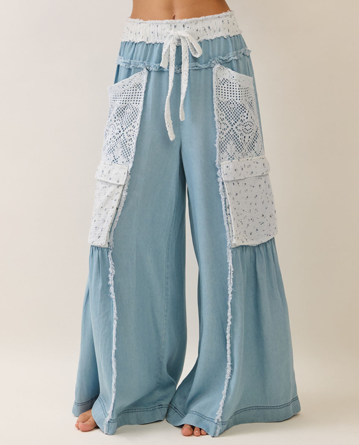 Light blue wide-leg pants with white crochet lace panel details on the front and pockets, featuring a white drawstring waistband, shown on a model against a plain beige background.