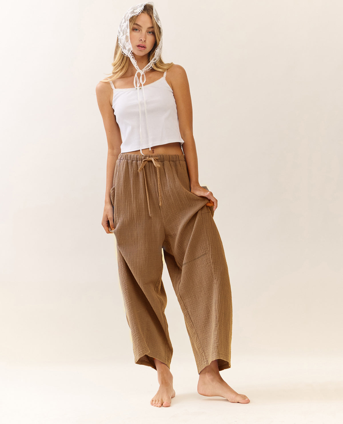 A woman stands barefoot against a plain warm beige studio background, wearing loose brown drawstring pants, a white cropped tank top, and a white patterned headscarf.