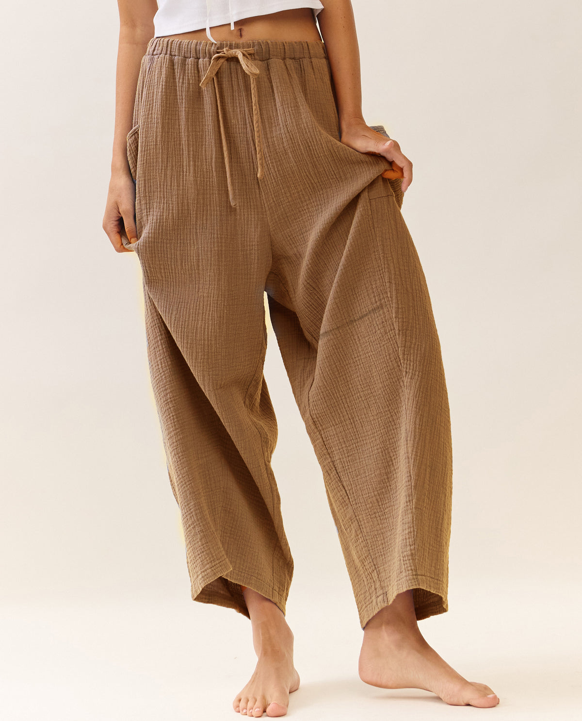 A person wearing loose, wide-leg cropped pants in a warm taupe-brown color with a matching drawstring waist, shown against a plain light beige studio background.