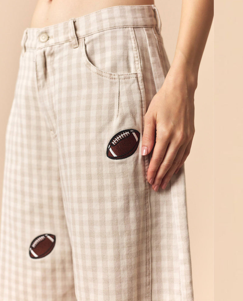 Close-up of a person wearing beige and white gingham pants with two brown football patch details on the legs, shown against a plain beige background.