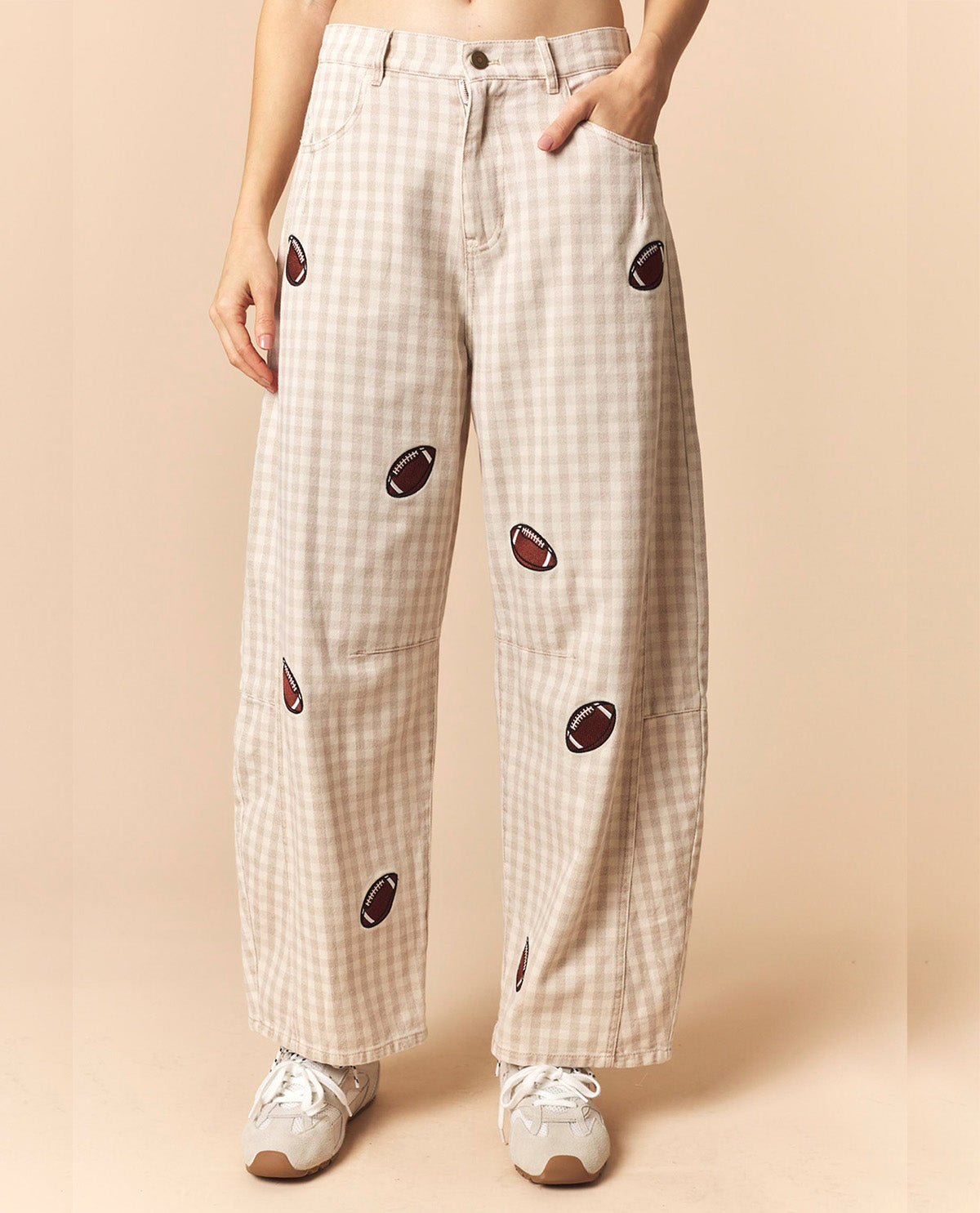 A person wearing beige and white gingham wide-leg pants with embroidered football motifs, paired with white sneakers, against a solid beige background.