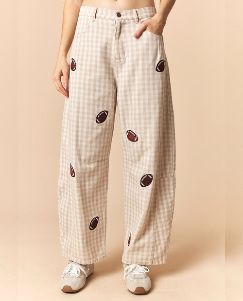 A person wearing beige and white gingham wide-leg pants with embroidered football motifs, paired with white sneakers, against a solid beige background.