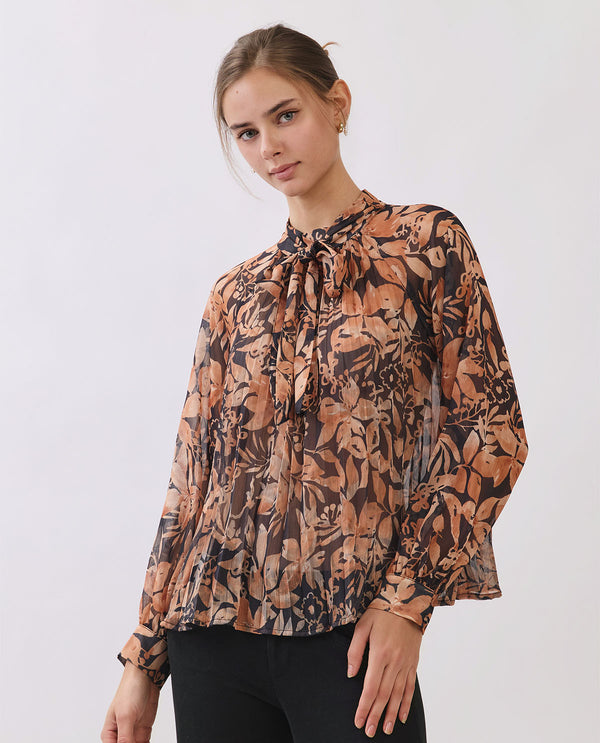 A woman wearing a long-sleeve blouse with a brown, beige, and black botanical leaf pattern, paired with black pants, photographed against a light gray studio background.