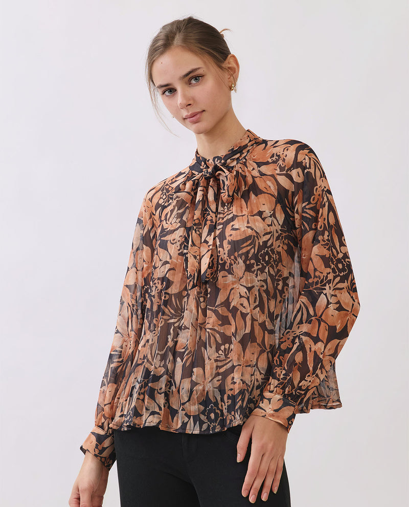 A woman wearing a long-sleeve blouse with a brown, beige, and black botanical leaf pattern, paired with black pants, photographed against a light gray studio background.