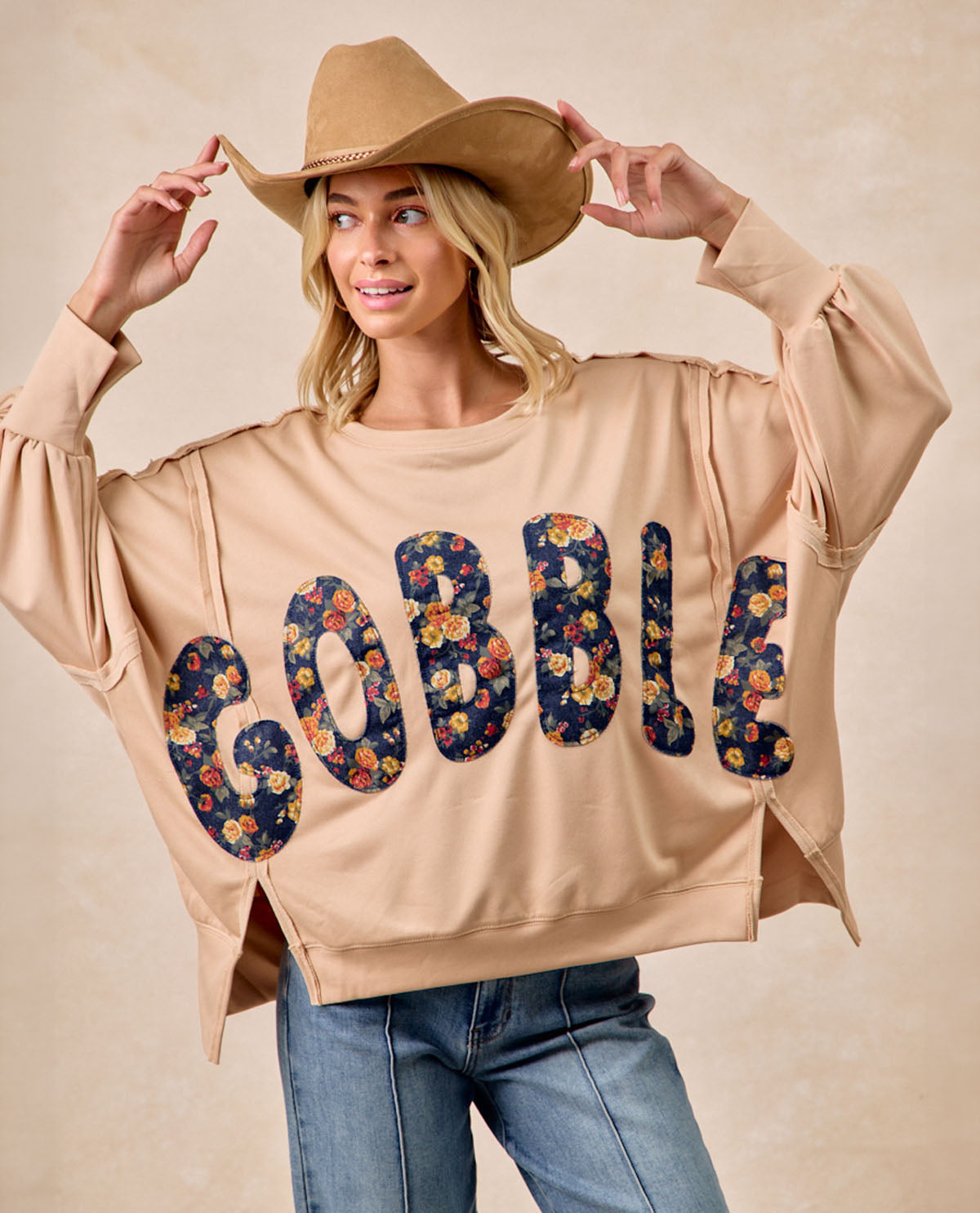 A woman wearing a beige cropped sweatshirt with the word 'GOBBLE' in large floral applique letters, paired with blue jeans and a tan cowboy hat, against a neutral beige studio background.