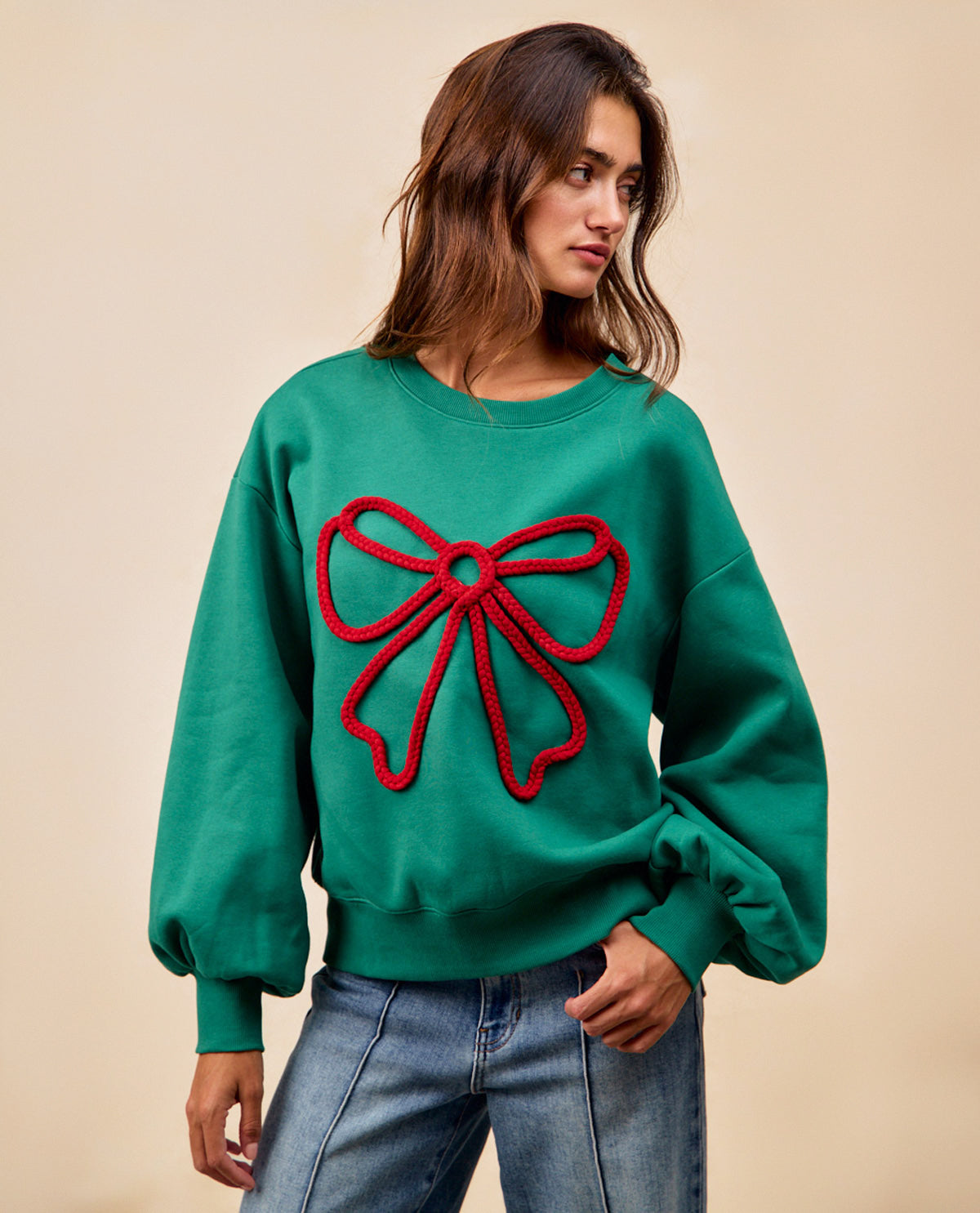 A woman wearing a green crewneck sweatshirt with a large red bow applique on the front, paired with blue jeans, set against a plain beige background.