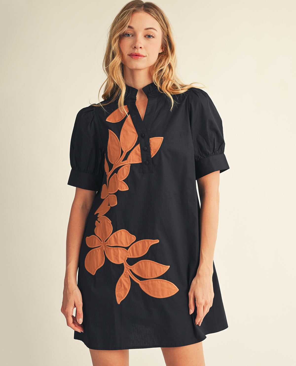 A woman modeling a black short-sleeve mini dress with a keyhole neckline and a large brown leaf pattern on the front, set against a plain beige background.