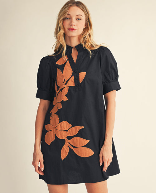 A woman modeling a black short-sleeve mini dress with a keyhole neckline and a large brown leaf pattern on the front, set against a plain beige background.