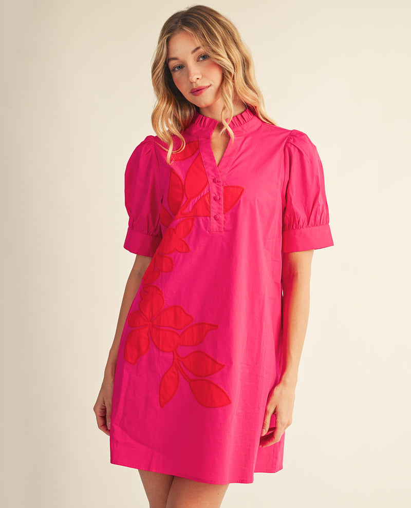 A woman modeling a short-sleeve bright pink dress with red floral embroidery and a buttoned collar, set against a plain beige studio background.