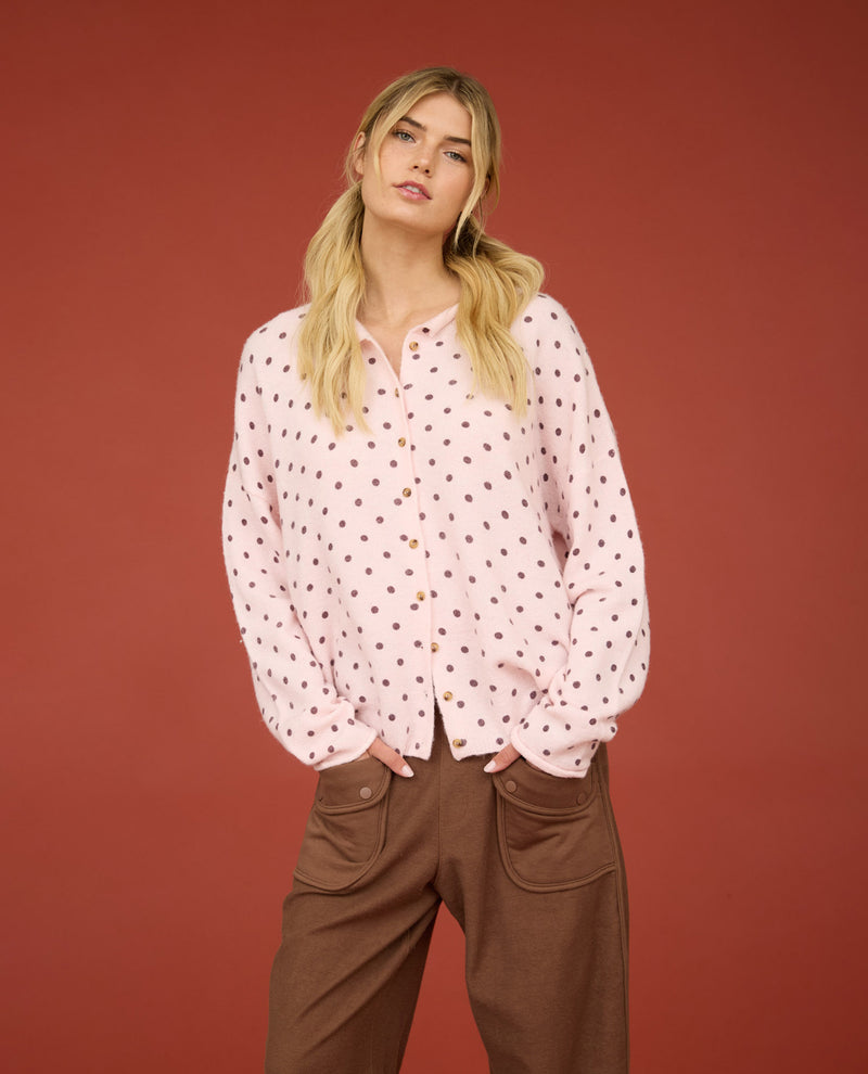 A blonde woman models a light pink long-sleeve button-up shirt with a small brown polka dot pattern, paired with brown pants, set against a solid terracotta red studio background.