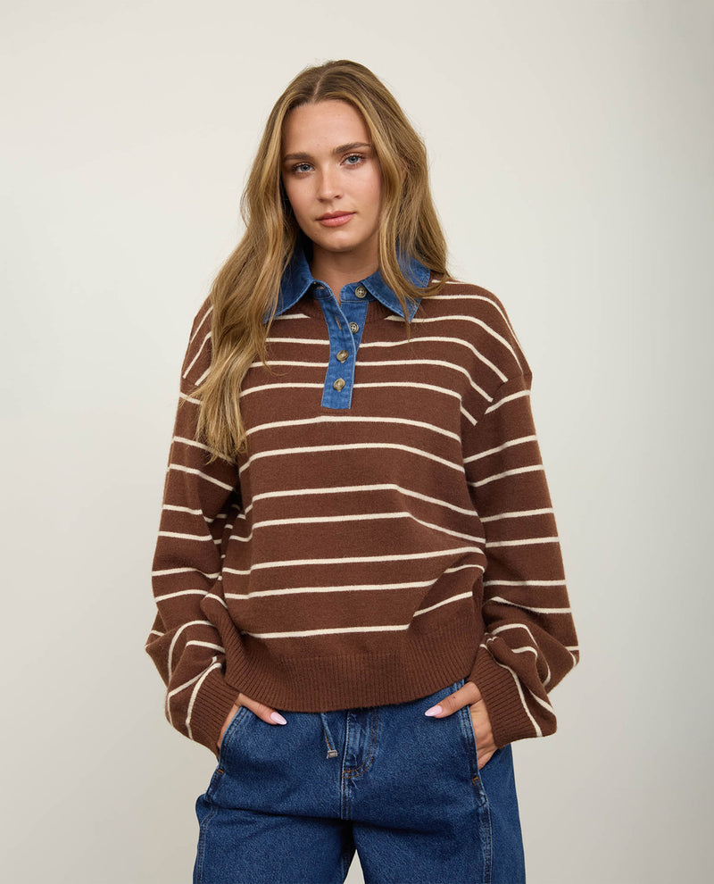 A woman modeling a brown and cream striped knit sweater with a blue denim collar and button placket, paired with blue jeans, against a plain light gray studio background.