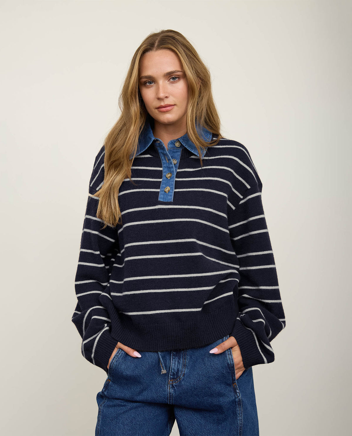 A woman modeling a navy blue and white horizontally striped knit polo sweater with a blue denim-style collar and button placket, paired with blue jeans, against a plain light beige studio background.