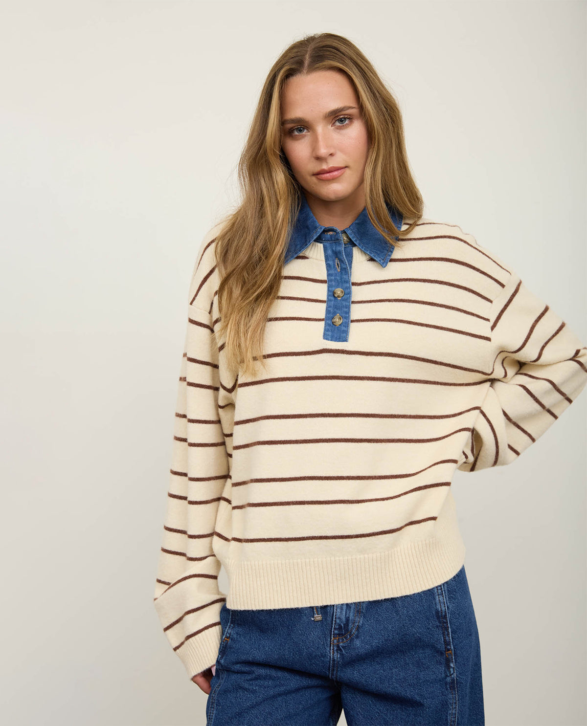 A woman modeling a cream and brown horizontally striped knit sweater with a blue denim polo collar and button placket, paired with blue jeans against a plain light background.