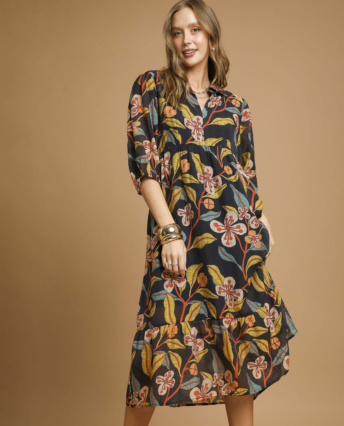 A woman models a black midi shirt dress with a vibrant floral print in orange, yellow, and white, set against a solid tan studio background.