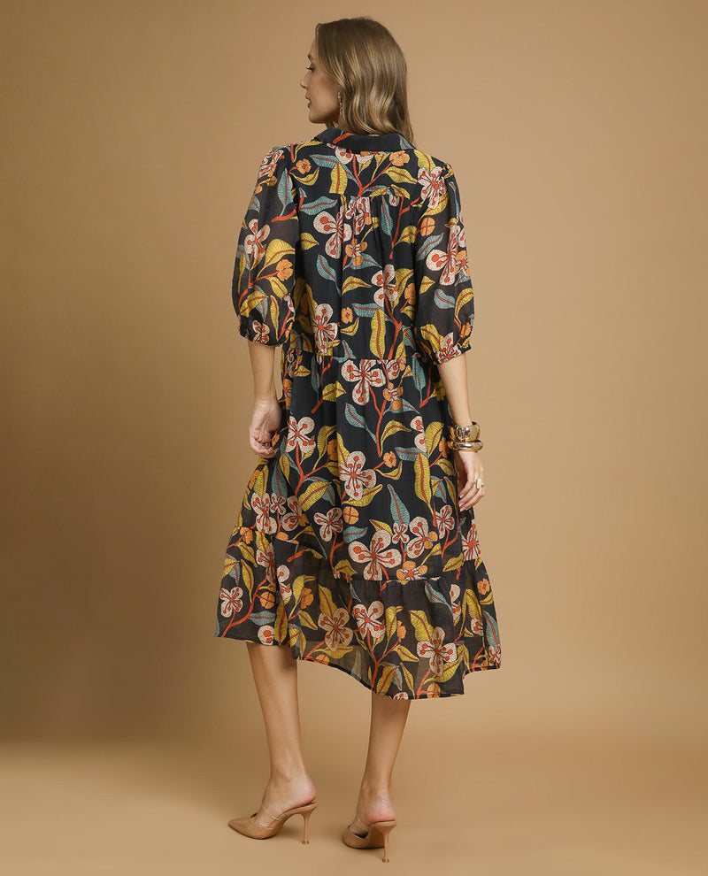 A woman shown from the back wearing a midi-length floral shirt dress with puff sleeves, featuring a black base with yellow, orange, and green botanical patterns, set against a solid tan studio background.