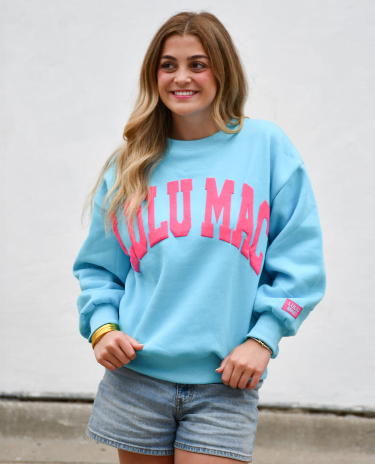 A woman wearing a light blue crewneck sweatshirt with 'LULU MAC' printed in pink across the chest, paired with denim shorts, standing against a plain white wall.