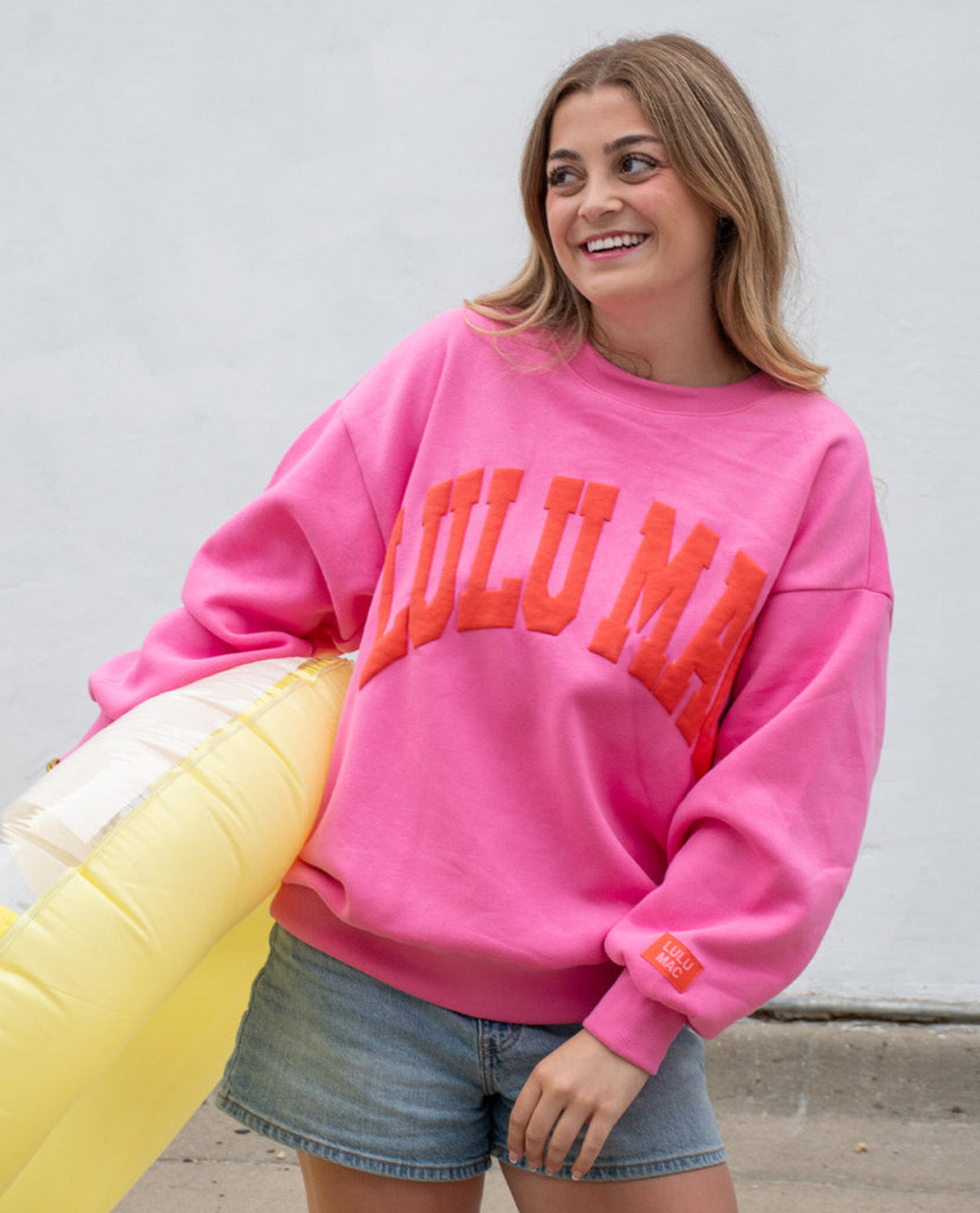 A woman wearing a bright pink crewneck sweatshirt with 'LULU MAC' printed in orange across the chest, paired with denim shorts and holding a yellow inflatable pool float against a plain light gray wall.