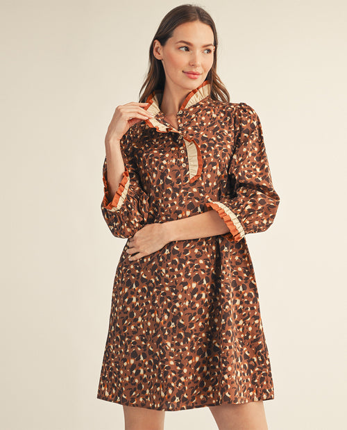 A woman modeling a brown leopard-print mini dress with orange and cream striped ruffled trim on the collar, cuffs, and placket, set against a plain beige studio background.
