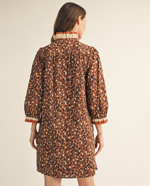 A woman shown from the back wearing a brown and black abstract patterned dress with beige and orange striped ruffled trim at the collar and cuffs, against a plain light beige studio background.