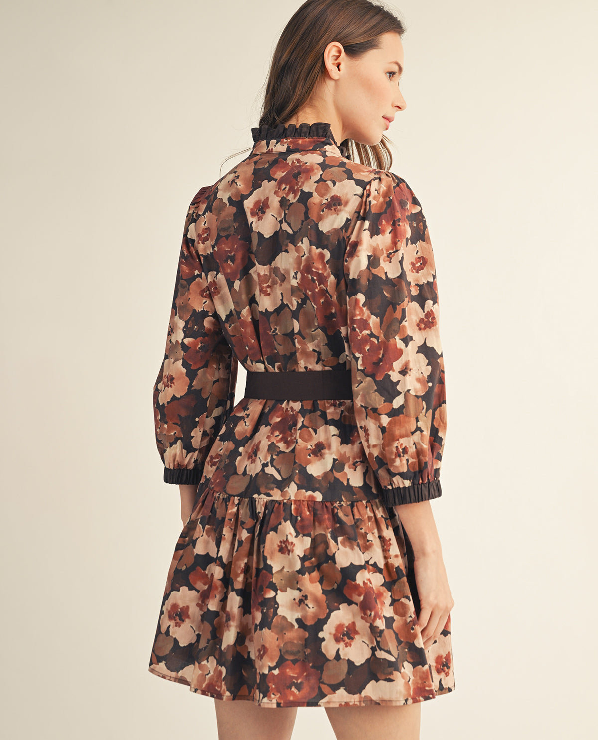 A woman modeling a short floral dress from the back, featuring a dark brown base with large beige, rust, and brown floral print, a dark brown waist belt, and ruffled trim at the collar and cuffs, set against a plain beige studio background.