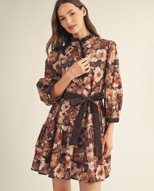 A woman modeling a long-sleeve floral mini dress with a dark brown waist tie, photographed against a plain light beige studio background.