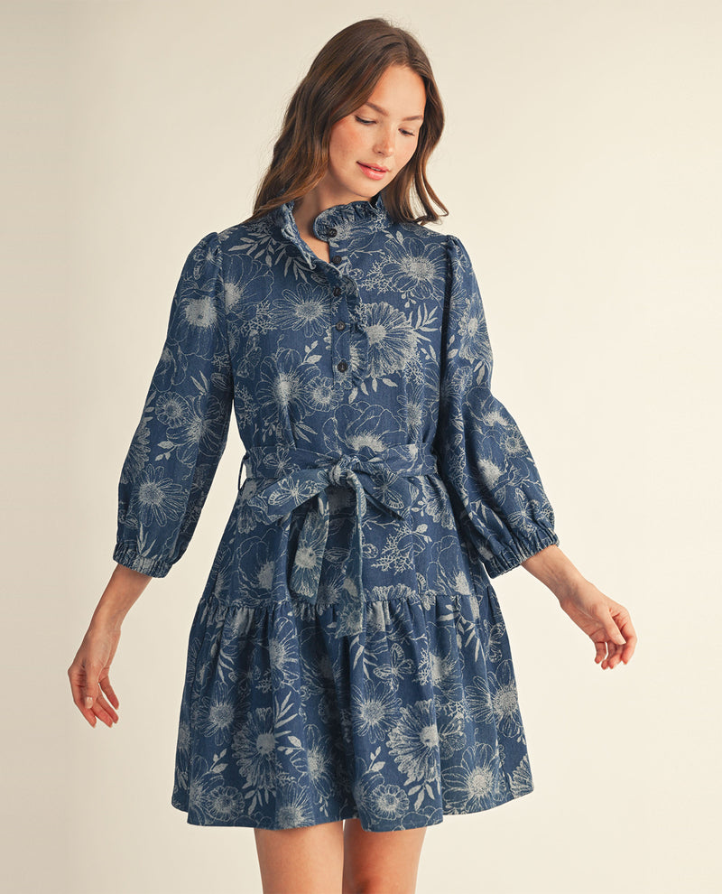 A woman wearing a blue floral long-sleeve mini dress with a tie waist, photographed against a plain light beige studio background.