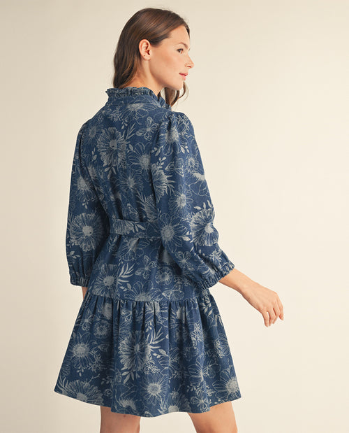A woman wearing a blue floral long-sleeve mini dress, shown from a side-back angle against a plain beige studio background.