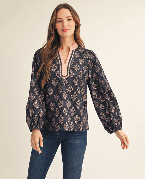 A woman modeling a long-sleeve navy blue blouse with a beige and white floral pattern, featuring a split neckline with pink and white trim, paired with dark blue jeans against a light beige background.